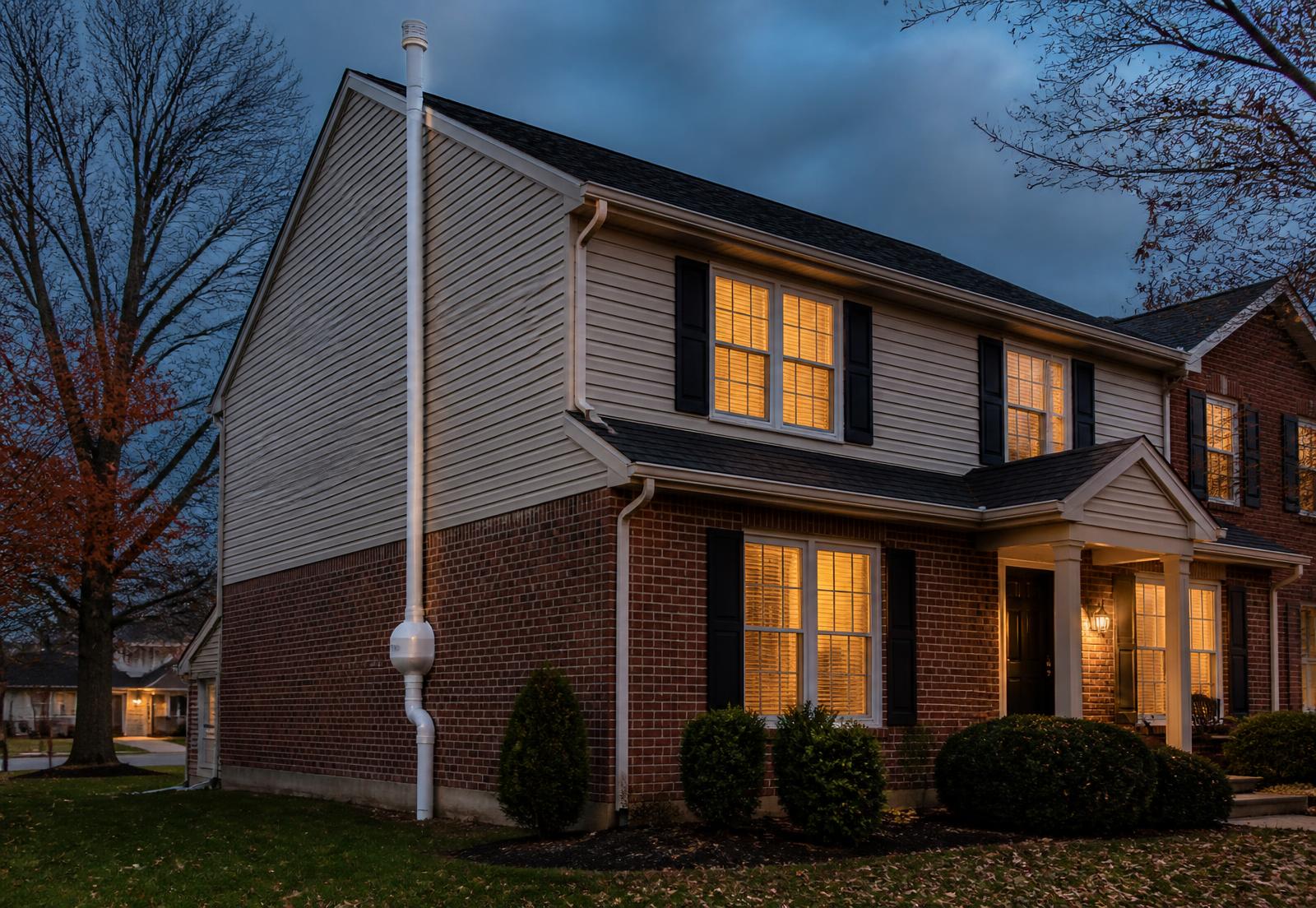 Indianapolis two-story brick home at dusk with a radon mitigation vent pipe routed up the exterior wall above the roofline